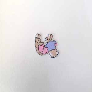 May include: Embroidered patch featuring two brown rabbits, one wearing a pink dress and the other a blue shirt.