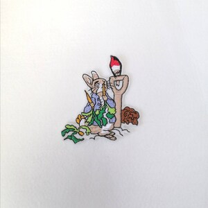 May include: An embroidered patch depicting Peter Rabbit holding a carrot, with a red robin perched on a spade. The patch is surrounded by green leaves and a brown mound of earth.