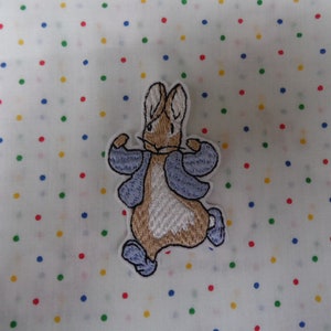 May include: Embroidered patch featuring Peter Rabbit from Beatrix Potter's children's book. The patch shows Peter Rabbit wearing a blue jacket and running with his arms outstretched.