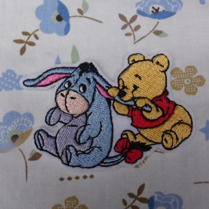 May include: Embroidered patch featuring Winnie the Pooh and Eeyore from the Winnie the Pooh cartoon. Winnie the Pooh is holding a red and white striped stick and Eeyore is sitting with a sad expression.