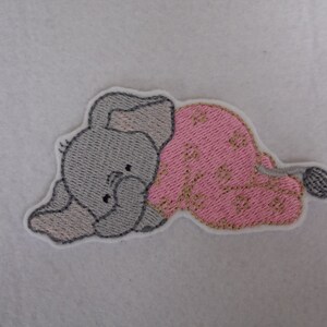 May include: Embroidered iron-on patch of a grey elephant wearing pink and gold patterned pyjamas. The elephant is lying down with its head resting on its front leg.