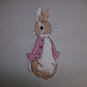 May include: Embroidered patch of Peter Rabbit wearing a pink jacket. The patch is white, brown, and pink.