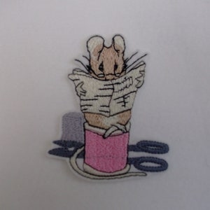 May include: Embroidered iron-on patch featuring a mouse reading a newspaper while sitting on a pink spool of thread with scissors nearby.