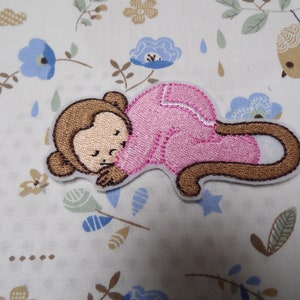 May include: Embroidered iron-on patch of a brown monkey wearing pink pyjamas and sleeping. The monkey's tail is curled around its body.