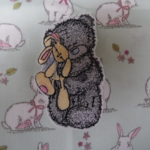 May include: An embroidered patch featuring a grey and white teddy bear holding a brown bunny rabbit. The bear is sitting upright and the bunny is in its arms. The patch is on a green background with white rabbits.