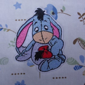 May include: Embroidered iron-on patch featuring a blue donkey character holding a red bag. The donkey has pink ears and a black nose.