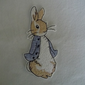 May include: Embroidered patch of a brown and white rabbit wearing a blue jacket. The rabbit has long ears and a white fluffy tail.