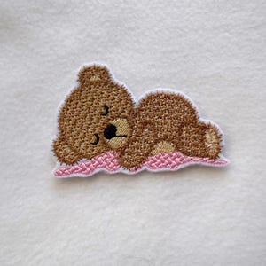 May include: An embroidered patch featuring a brown teddy bear lying on a pink pillow, with a white border. The bear has a black nose and eyes. Suitable for sewing or ironing onto fabric.