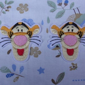 May include: Two embroidered iron-on patches featuring the face of the cartoon character Tigger from Winnie the Pooh. The patches are orange, yellow, black, white, and pink.