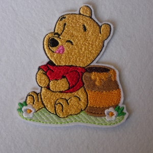 May include: Embroidered iron-on patch featuring Winnie the Pooh sitting with a honey pot. The patch is yellow, red, brown, and orange with green and white accents.