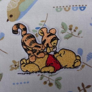 May include: Embroidered iron-on patch featuring Winnie the Pooh and Tigger. Winnie the Pooh is lying down with his eyes closed, while Tigger is standing on his back and looking at Winnie the Pooh.