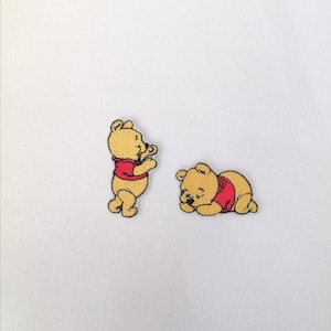 May include: Two embroidered iron-on patches featuring Winnie the Pooh. One patch shows Winnie standing and the other shows him sleeping.