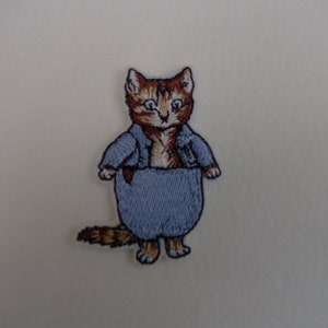 May include: Iron-on patch featuring an embroidered brown and white cat wearing a blue jacket and blue trousers. The cat has a white face and brown eyes.