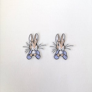May include: Two iron-on patches featuring Peter Rabbit in a blue jacket, with white accents, and brown fur.