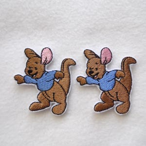 May include: Two embroidered patches of a cartoon kangaroo wearing a blue shirt. The kangaroo is brown with pink ears and a white outline. These patches are designed for application on clothing or accessories.