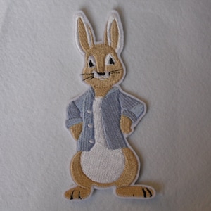 May include: Embroidered iron-on patch featuring a brown rabbit wearing a blue denim jacket. The rabbit has white fur on its belly and a white tail.