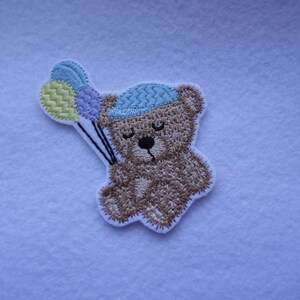 May include: Embroidered patch of a brown teddy bear wearing a blue hat and holding three balloons in yellow, blue, and purple.