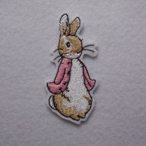 May include: Iron-on patch featuring Peter Rabbit wearing a pink jacket. The patch is white with brown and gold accents.