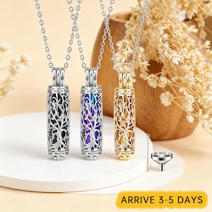 May include: Three cylindrical pendant necklaces featuring an intricate tree of life cutout design. The pendants come in silver, iridescent blue, and gold tones, each suspended on a matching chain. A small metal funnel and pin are included.