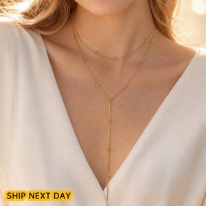 May include: A gold-toned layered necklace featuring a delicate chain with small, evenly spaced spherical beads. The design includes a shorter upper strand and a longer lower strand that forms a Y-shape, resting against a white fabric garment.
