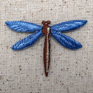 May include: A blue embroidered dragonfly with brown accents on a beige fabric background.