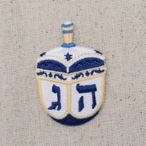 Può includere: Un dreidel ricamato blu e bianco con le lettere ebraiche 'nun', 'guimel', 'hei' e 'shin' sui lati. Il dreidel ha una stella dorata al centro.