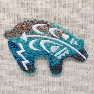 Pode incluir: Um remendo bordado com um urso azul turquesa e marrom com desenhos geométricos brancos. O urso está olhando para a direita e tem um design de pena estilizado nas costas.