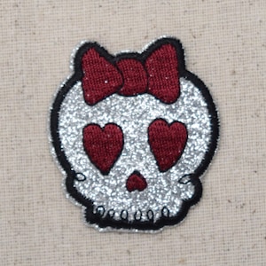 May include: A silver glitter skull with red heart eyes and a red bow on top.
