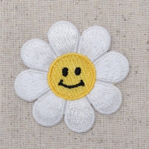 Pode incluir: Flor de margarida branca com um rosto sorridente amarelo no centro. A flor é bordada em um tecido bege.