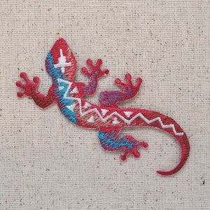 May include: Red and blue embroidered gecko with a geometric pattern. The gecko is on a beige fabric background.