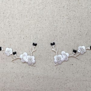 May include: Two embroidered floral patches featuring white blossoms with silver detailing and black accents. The patches are arranged in a branch-like design, perfect for adding a decorative touch to clothing or accessories.