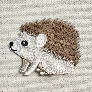 以下が含まれることがあります： 茶色の背中と白いお腹を持つハリネズミの刺繍パッチ。ハリネズミは左を向いており、小さな黒い鼻と目があります。