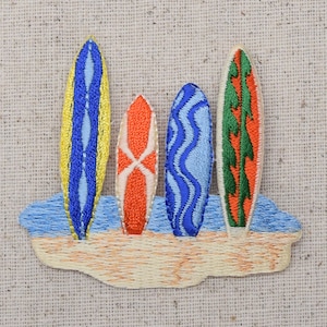Może przedstawiać: Naszywka haftowana z czterema kolorowymi deskami surfingowymi na beżowym tle z tkaniny. Deski surfingowe są niebieskie, żółte, pomarańczowe i zielone.