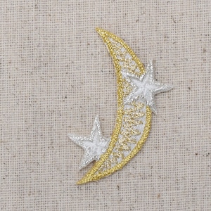 May include: A gold crescent moon with a silver star on each side. The moon and stars are embroidered on a beige fabric.
