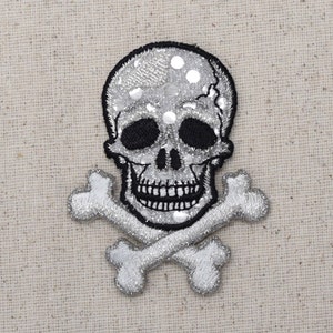 Peut inclure: Un écusson en forme de crâne et d'os croisés en paillettes argentées. Le crâne est noir et blanc avec un sourire narquois. Les os croisés sont en paillettes argentées.