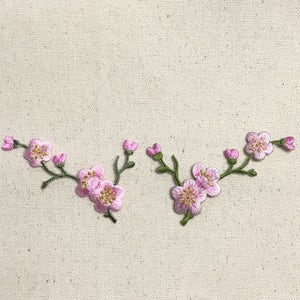 May include: Two embroidered pink flower branches with green stems and leaves on a white fabric background.