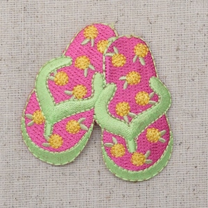 May include: A pair of pink flip-flops with yellow and green embroidered details. The flip-flops are on a beige fabric background.