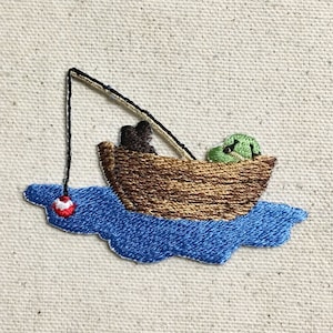 Op de afbeelding: Gestikt patch van een bruin bootje met een groene kikker erin, een hengel met een rode dobber en blauw water.