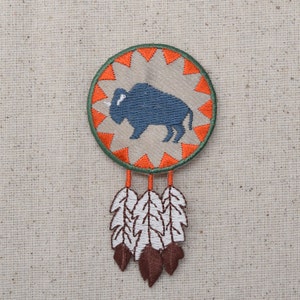May include: A round, embroidered patch with a blue buffalo silhouette inside a circle of orange and green. The patch has three brown and white feathers hanging from the bottom.