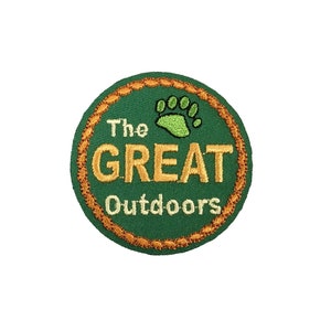 Pode incluir: Etiqueta bordada verde com borda de corda dourada. A etiqueta apresenta uma pegada verde e o texto "The GREAT Outdoors".