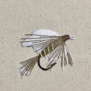 Könnte beinhalten: Gestickte Fliegenfischer-Köder in Weiß, Braun und Grün. Die Fliege ist auf einen beigefarbenen Stoff gestickt, der die komplizierten Details des Designs zeigt. Der Haken ist dunkelbraun.