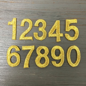 May include: Ten gold embroidered iron-on numbers, 1 through 0, on a wood background.