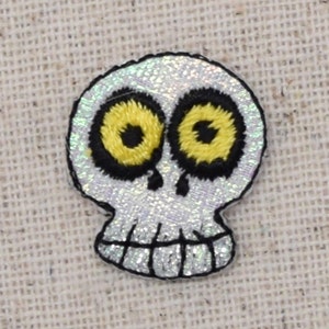 Puede incluir: Un cráneo blanco y plateado brillante con contornos negros y ojos amarillos. El cráneo está bordado en una tela blanca.