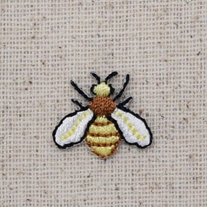 May include: A small embroidered bee with yellow and brown stripes and white wings on a beige fabric background.