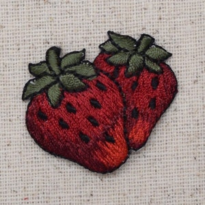 Peut inclure: Deux fraises brodées en rouge et vert sur un fond de tissu beige.