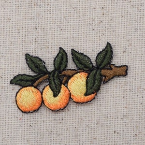 Puede incluir: Parche bordado de una pequeña rama de naranjo con tres naranjas. La rama es marrón con hojas verdes y las naranjas son de color naranja claro.