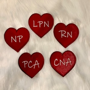 May include: Five red felt heart-shaped patches with white embroidered text. The text on the patches reads: LPN, RN, NP, PCA, and CNA.