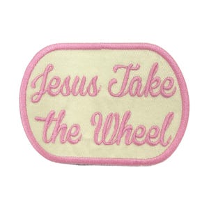 May include: Embroidered patch with the phrase "Jesus Take the Wheel" in pink script lettering. The patch has a cream-colored background and a pink border, with an oval shape.