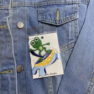 Puede incluir: Chaqueta vaquera con un parche de rana. El parche presenta una rana verde surfeando sobre una tabla de surf amarilla y azul. El parche está adherido a una funda de plástico transparente con el texto "Pie Patches".