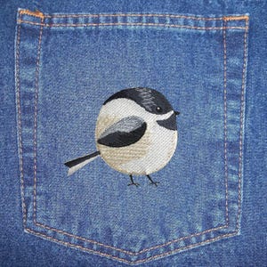 May include: Embroidered black and white chickadee bird on a blue denim pocket.